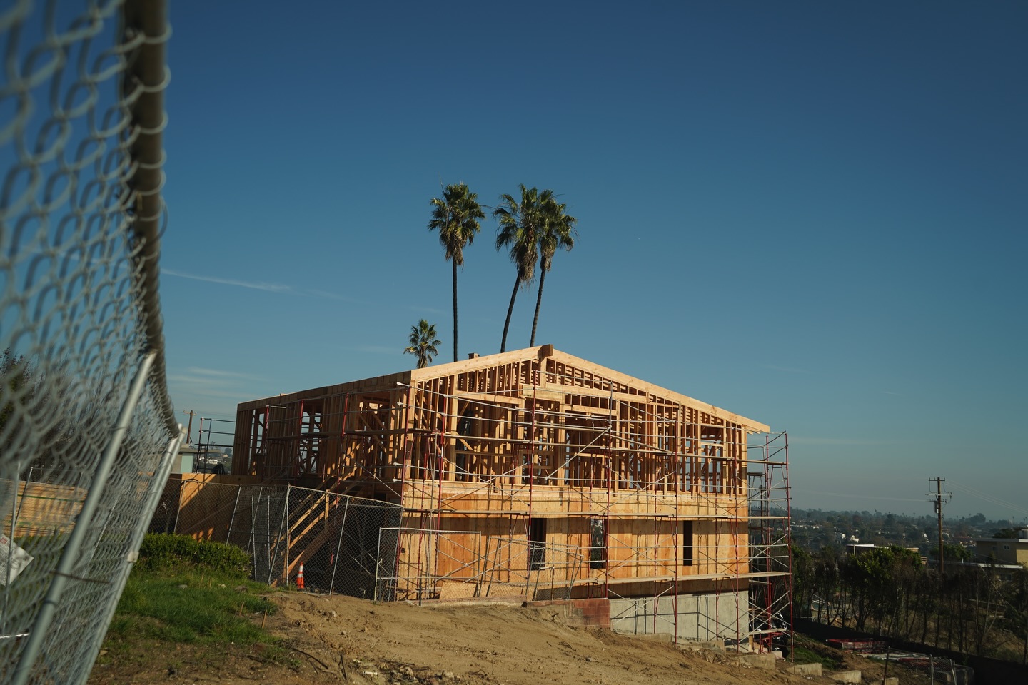 Two-story wood frame house wrapped in scaffolding on a hillside, palm trees behind, seen through a chain-link fence