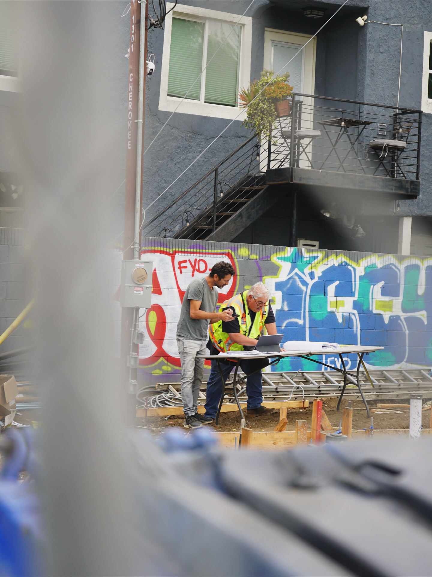 Two men reviewing plans on a laptop at a folding table on a job site, graffiti wall behind them