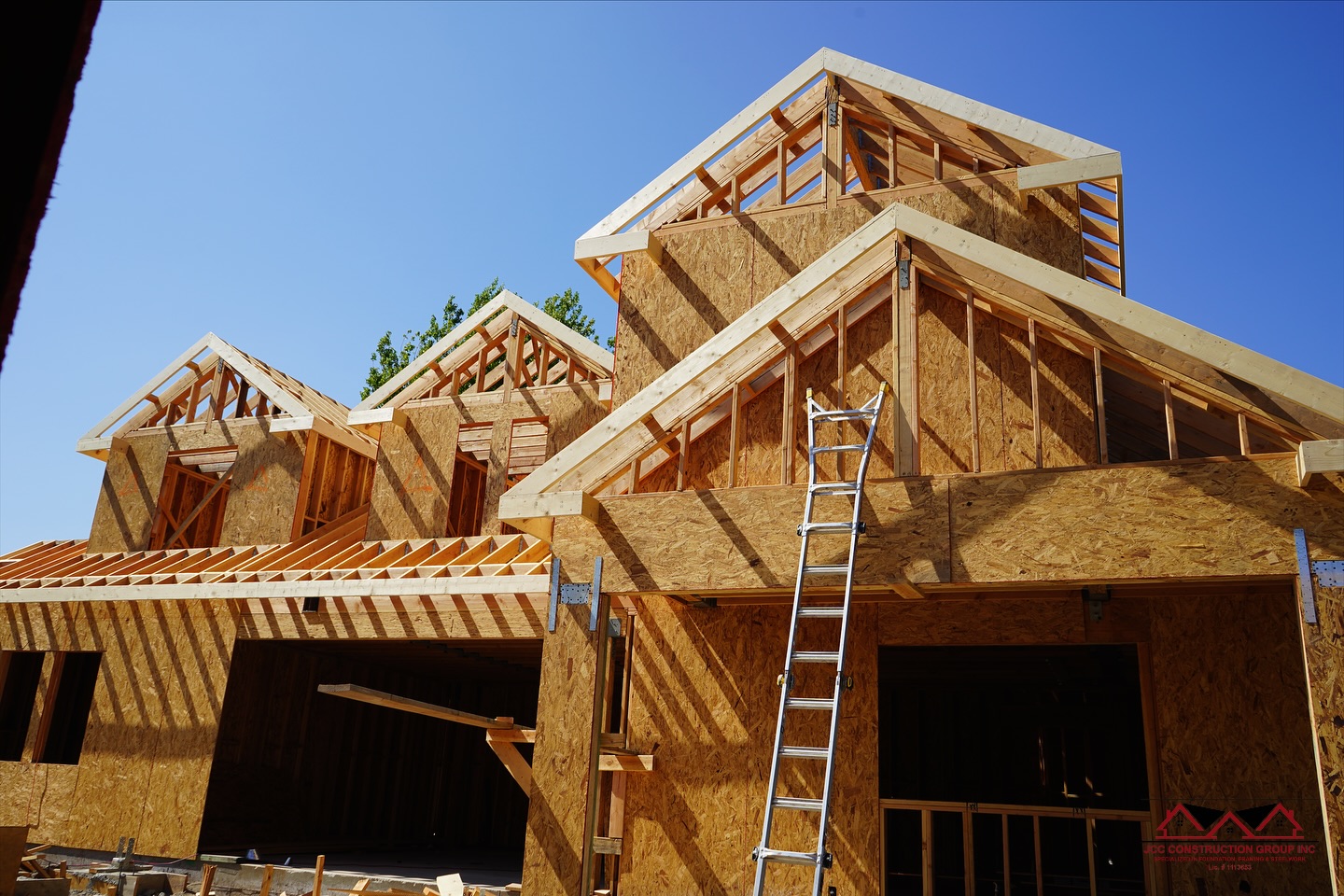 Framed roofline with dormers and OSB sheathing against a blue sky, ladder leaning on the wall